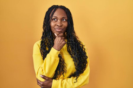 Photo for African woman standing over yellow background looking confident at the camera with smile with crossed arms and hand raised on chin. thinking positive. - Royalty Free Image