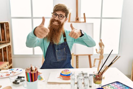 Photo for Redhead man with long beard painting clay bowl at art studio approving doing positive gesture with hand, thumbs up smiling and happy for success. winner gesture. - Royalty Free Image