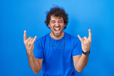 Photo for Hispanic young man standing over blue background shouting with crazy expression doing rock symbol with hands up. music star. heavy music concept. - Royalty Free Image
