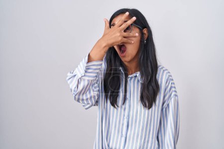 Foto de Mujer hispana joven con anteojos asomándose en shock cubriendo cara y ojos con la mano, mirando a través de los dedos con expresión avergonzada. - Imagen libre de derechos