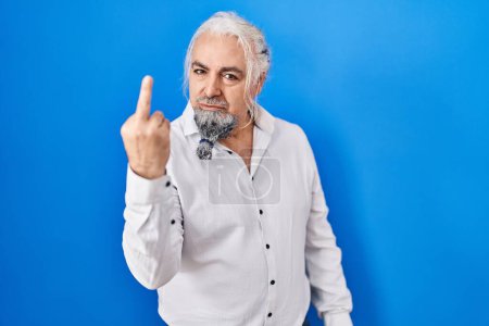 Foto de Middle age man with grey hair standing over blue background showing middle finger, impolite and rude fuck off expression - Imagen libre de derechos