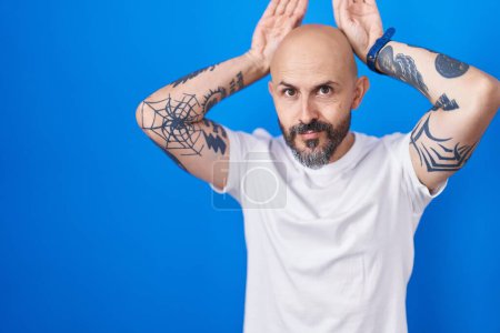 Foto de Hombre hispano con tatuajes de pie sobre fondo azul haciendo un gesto de orejas de conejo con palmas de manos que parecen cínicas y escépticas. concepto de conejo de Pascua. - Imagen libre de derechos