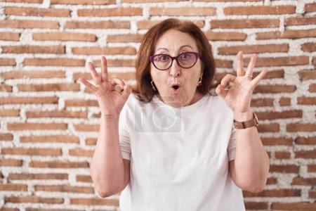 Foto de Senior woman with glasses standing over bricks wall looking surprised and shocked doing ok approval symbol with fingers. crazy expression - Imagen libre de derechos