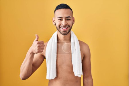 Téléchargez les photos : Jeune homme hispanique debout torse nu avec une serviette faisant des pouces heureux geste avec la main. approuvant l'expression regardant la caméra montrant le succès. - en image libre de droit