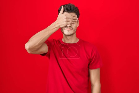 Foto de Young hispanic man standing over red background covering eyes with hand, looking serious and sad. sightless, hiding and rejection concept - Imagen libre de derechos