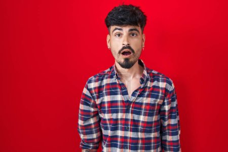 Photo for Young hispanic man with beard standing over red background afraid and shocked with surprise and amazed expression, fear and excited face. - Royalty Free Image