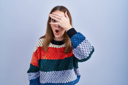 Photo for Young hispanic girl standing over blue background peeking in shock covering face and eyes with hand, looking through fingers with embarrassed expression. - Royalty Free Image