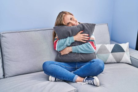 Téléchargez les photos : Young blonde woman hugging cushion sitting on sofa at home - en image libre de droit