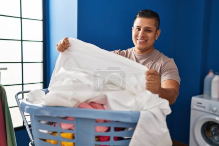 Foto de Joven latino sonriendo confiado colgando ropa en tendedero en la lavandería - Imagen libre de derechos