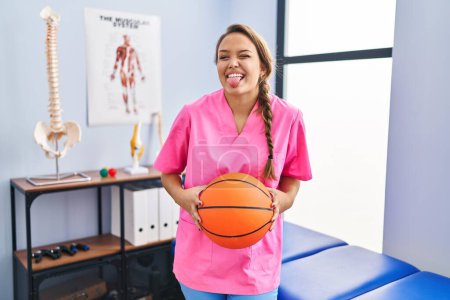 Photo for Young hispanic woman working at physiotherapy clinic holding basketball ball sticking tongue out happy with funny expression. - Royalty Free Image