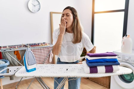 Foto de Young hispanic woman ironing clothes at laundry room bored yawning tired covering mouth with hand. restless and sleepiness. - Imagen libre de derechos