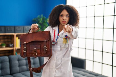 Foto de Young african american doctor woman holding bag for home assistance pointing with finger to the camera and to you, confident gesture looking serious - Imagen libre de derechos