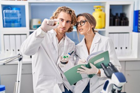 Foto de Man and woman wearing scientist uniform holding pills writing on notebook at laboratory - Imagen libre de derechos