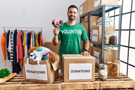 Foto de Young hispanic man wearing volunteer uniform packing donations box at charity center - Imagen libre de derechos