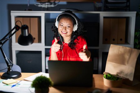 Téléchargez les photos : Jeune femme asiatique travaillant au bureau avec ordinateur portable la nuit approuvant faire un geste positif avec la main, pouces levés souriant et heureux pour le succès. geste gagnant. - en image libre de droit