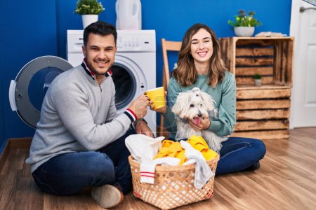 Photo for Man and woman drinking coffee washing clothes with dog at laundry room - Royalty Free Image