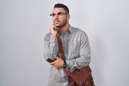 Foto de Young hispanic man wearing suitcase with hand on chin thinking about question, pensive expression. smiling with thoughtful face. doubt concept. - Imagen libre de derechos