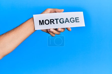 Foto de Hand of caucasian man holding paper with mortgage word over isolated blue background - Imagen libre de derechos