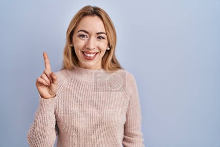Photo for Hispanic woman standing over blue background showing and pointing up with finger number one while smiling confident and happy. - Royalty Free Image