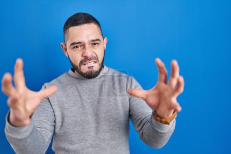 Photo for Hispanic man standing over blue background shouting frustrated with rage, hands trying to strangle, yelling mad - Royalty Free Image