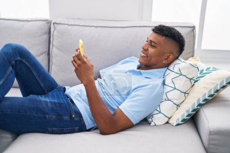 Photo for Young latin man using smartphone lying on sofa at home - Royalty Free Image