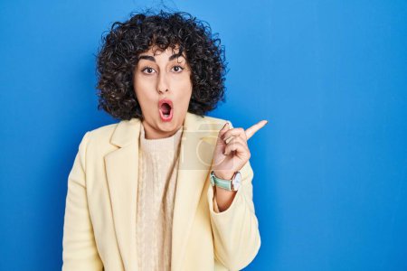 Photo for Young brunette woman with curly hair standing over blue background surprised pointing with finger to the side, open mouth amazed expression. - Royalty Free Image
