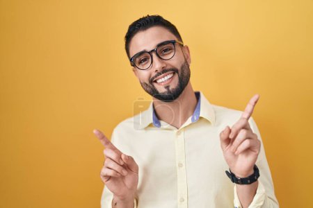 Foto de Joven hispano vestido con ropa de negocios y gafas sonriendo confiado señalando con los dedos a diferentes direcciones. espacio de copia para publicidad - Imagen libre de derechos