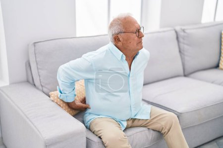 Foto de Hombre mayor de pelo gris que sufre de dolor de espalda sentado en el sofá en casa - Imagen libre de derechos