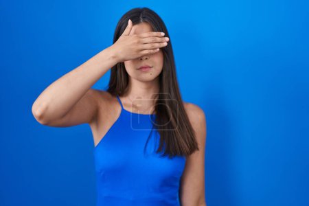 Photo for Hispanic woman standing over blue background covering eyes with hand, looking serious and sad. sightless, hiding and rejection concept - Royalty Free Image