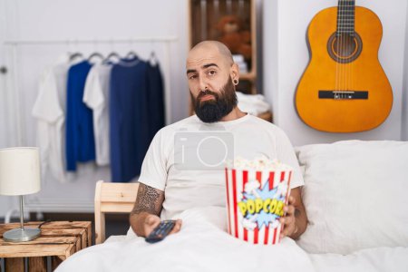 Foto de Joven hispano con barba y tatuajes comiendo palomitas de maíz en la cama deprimido y preocupado por la angustia, llorando enojado y asustado. expresión triste. - Imagen libre de derechos