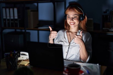 Photo for Young caucasian woman working at the office at night success sign doing positive gesture with hand, thumbs up smiling and happy. cheerful expression and winner gesture. - Royalty Free Image
