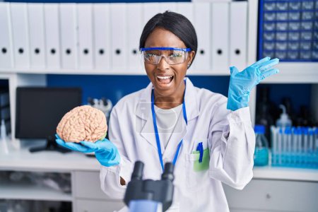 Téléchargez les photos : Jeune femme africaine travaillant au laboratoire scientifique tenant le cerveau célébrant la victoire avec un sourire heureux et l'expression gagnante avec les mains levées - en image libre de droit
