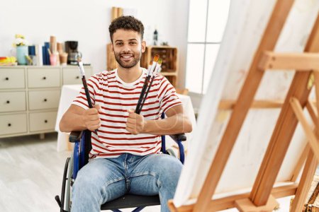 Téléchargez les photos : Jeune artiste arabe handicapé tenant des pinceaux assis en fauteuil roulant au studio d'art. - en image libre de droit