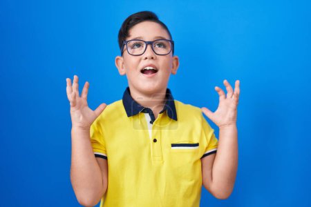 Téléchargez les photos : Young hispanic kid standing over blue background celebrating mad and crazy for success with arms raised and closed eyes screaming excited. winner concept - en image libre de droit
