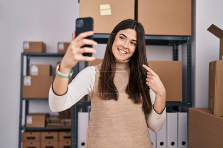Foto de Joven morena que trabaja en el comercio electrónico de pequeñas empresas tomando selfie sonriendo feliz señalando con la mano y el dedo - Imagen libre de derechos