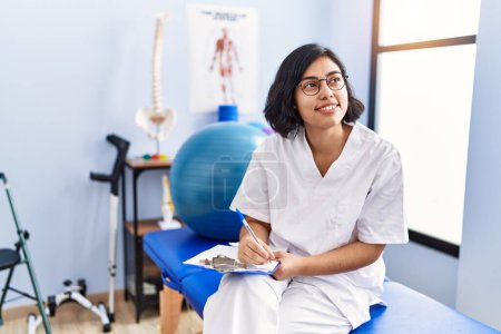 Photo for Young latin woman wearing physiotherapist uniform writing on clipboard at physiotherapy clinic - Royalty Free Image