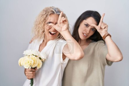 Foto de Madre e hija sosteniendo ramo de flores blancas burlándose de la gente con los dedos en la frente haciendo gesto perdedor burlándose e insultando. - Imagen libre de derechos