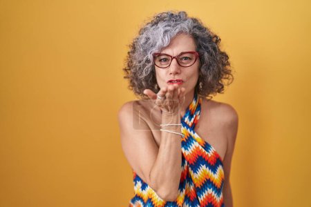 Photo for Middle age woman with grey hair standing over yellow background looking at the camera blowing a kiss with hand on air being lovely and sexy. love expression. - Royalty Free Image