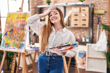 Foto de Mujer joven caucásica en el estudio de arte sosteniendo paleta estresada y frustrada con la mano en la cabeza, cara sorprendida y enojada - Imagen libre de derechos