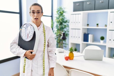 Photo for Young hispanic woman as nutritionist doctor holding weighing machine scared and amazed with open mouth for surprise, disbelief face - Royalty Free Image