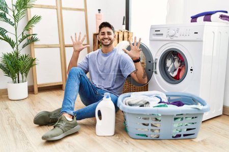 Photo for Young hispanic man putting dirty laundry into washing machine showing and pointing up with fingers number nine while smiling confident and happy. - Royalty Free Image