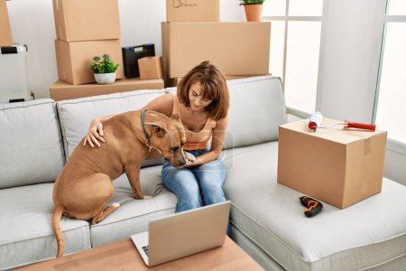 Téléchargez les photos : Jeune femme caucasienne utilisant ordinateur portable assis sur le canapé avec chien à la maison - en image libre de droit