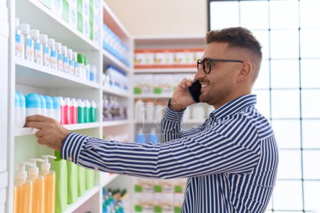 Photo for Young hispanic man customer talking on smartphone holding product on shelving at pharmacy - Royalty Free Image