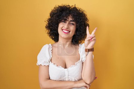 Téléchargez les photos : Young brunette woman with curly hair standing over yellow background smiling with happy face winking at the camera doing victory sign with fingers. number two. - en image libre de droit