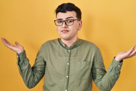 Non binary person standing over yellow background clueless and confused expression with arms and hands raised. doubt concept. 