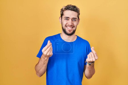 Téléchargez les photos : Homme hispanique avec barbe debout sur fond jaune faisant geste d'argent avec les mains, demandant le paiement du salaire, millionnaire entreprise - en image libre de droit
