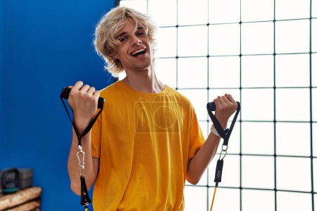 Téléchargez les photos : Jeune homme blond souriant confiant en utilisant l'entraînement de bande élastique au centre sportif - en image libre de droit