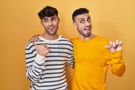 Foto de Joven pareja gay hispana de pie sobre fondo amarillo sonriendo con cara feliz mirando y señalando hacia un lado con el pulgar hacia arriba. - Imagen libre de derechos