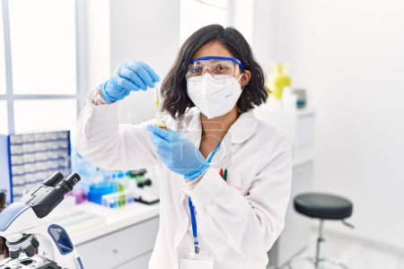 Foto de Young latin woman wearing scientist uniform and medical mask using pipette at laboratory - Imagen libre de derechos