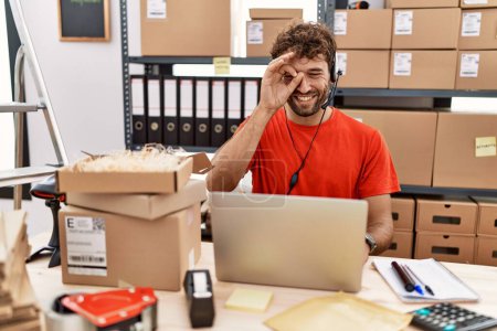 Téléchargez les photos : Young hispanic call center agent man working at warehouse doing ok gesture with hand smiling, eye looking through fingers with happy face. - en image libre de droit